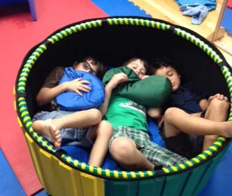 Three boys resting together in a round yellow and green pen with pillows